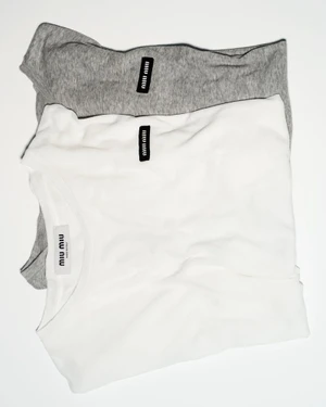 Two layered folded t-shirts, one white and one grey, featuring small black brand labels against a plain white background.