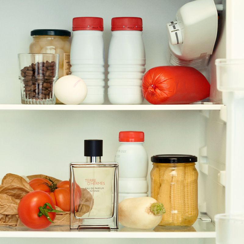Inside a refrigerator with food items, clear glass bottle of cologne, tomatoes, and various containers on white shelves.