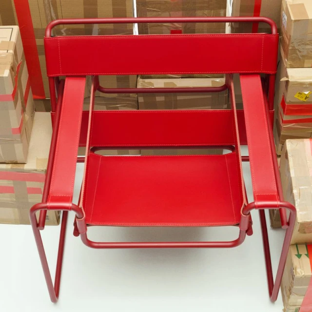 A red metal-framed chair with red leather straps is centered in a shot with blurred cardboard boxes filling the background.