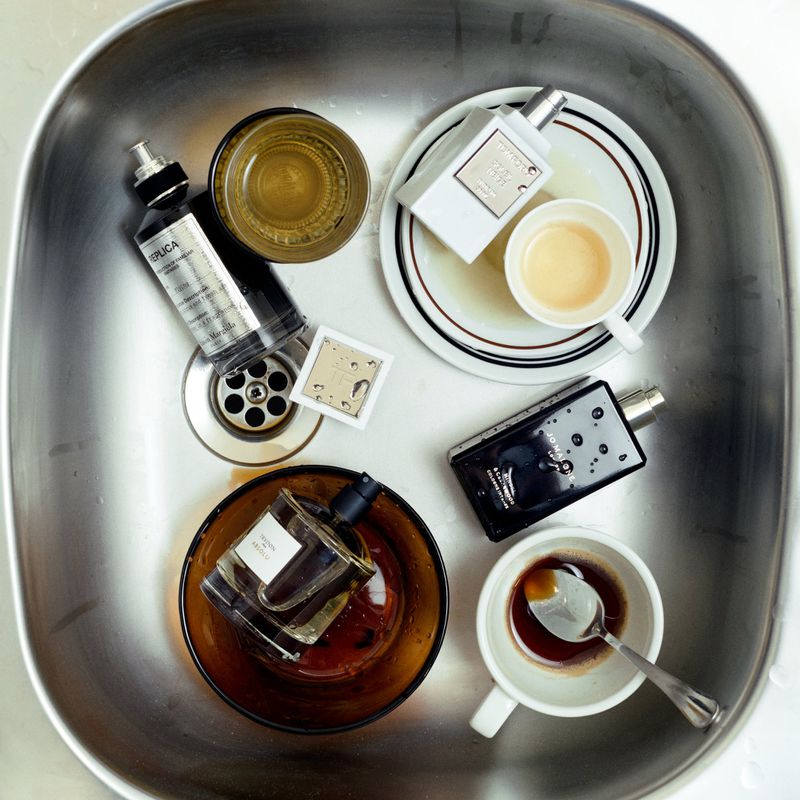 Overhead shot of fragrance bottles and cups in a stainless steel sink. Beauty product flat lay in a metallic setting.