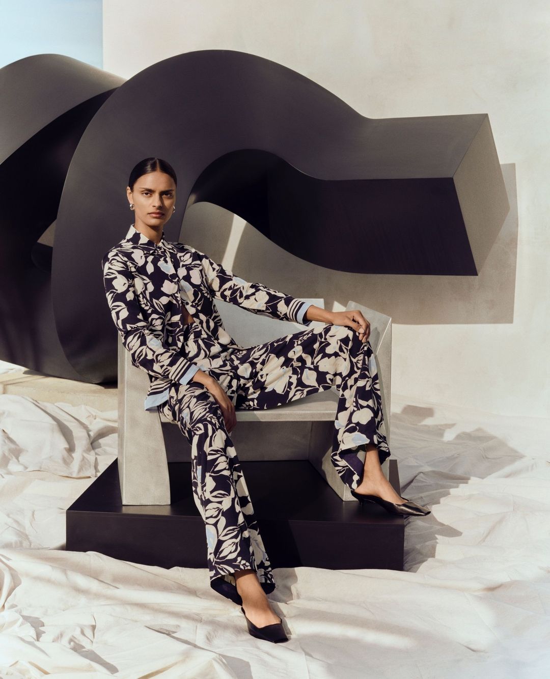 Fashion shot of a woman in patterned pajamas sitting on a chair in front of a black abstract sculpture in a studio setting.