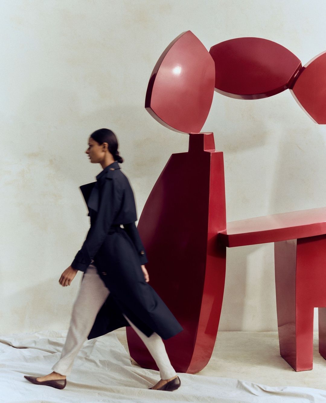 Fashion editorial image features a woman in a dark trench coat and light pants walking past a large red abstract sculpture indoors.