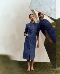Woman wearing a dark denim midi skirt and matching denim shirt standing near a geometric reflective sculpture on grass.
