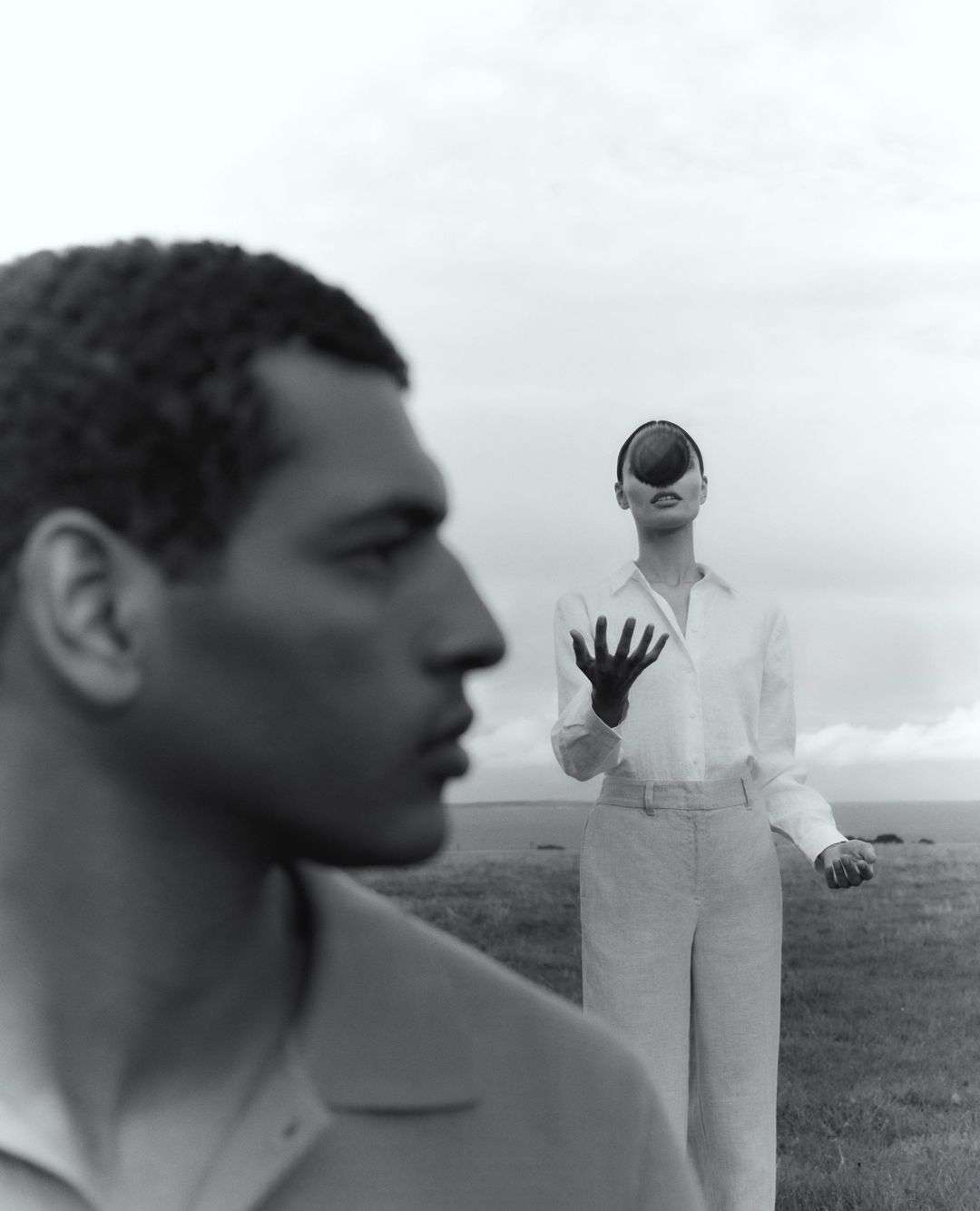 Monochrome portrait of a man in foreground profile and woman in background holding fruit to her face in an open grassy landscape.