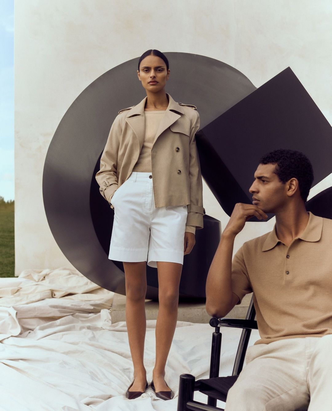 Two fashion models in outdoor setting. Woman stands in trench coat and shorts. Man sits in polo shirt, with sculpture in background.