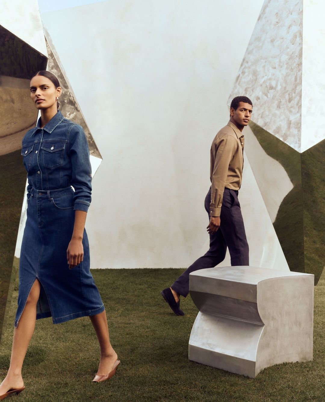 A woman in a denim dress and a man in a tan shirt walk in a modern outdoor space with green turf and geometric white architectural elements.