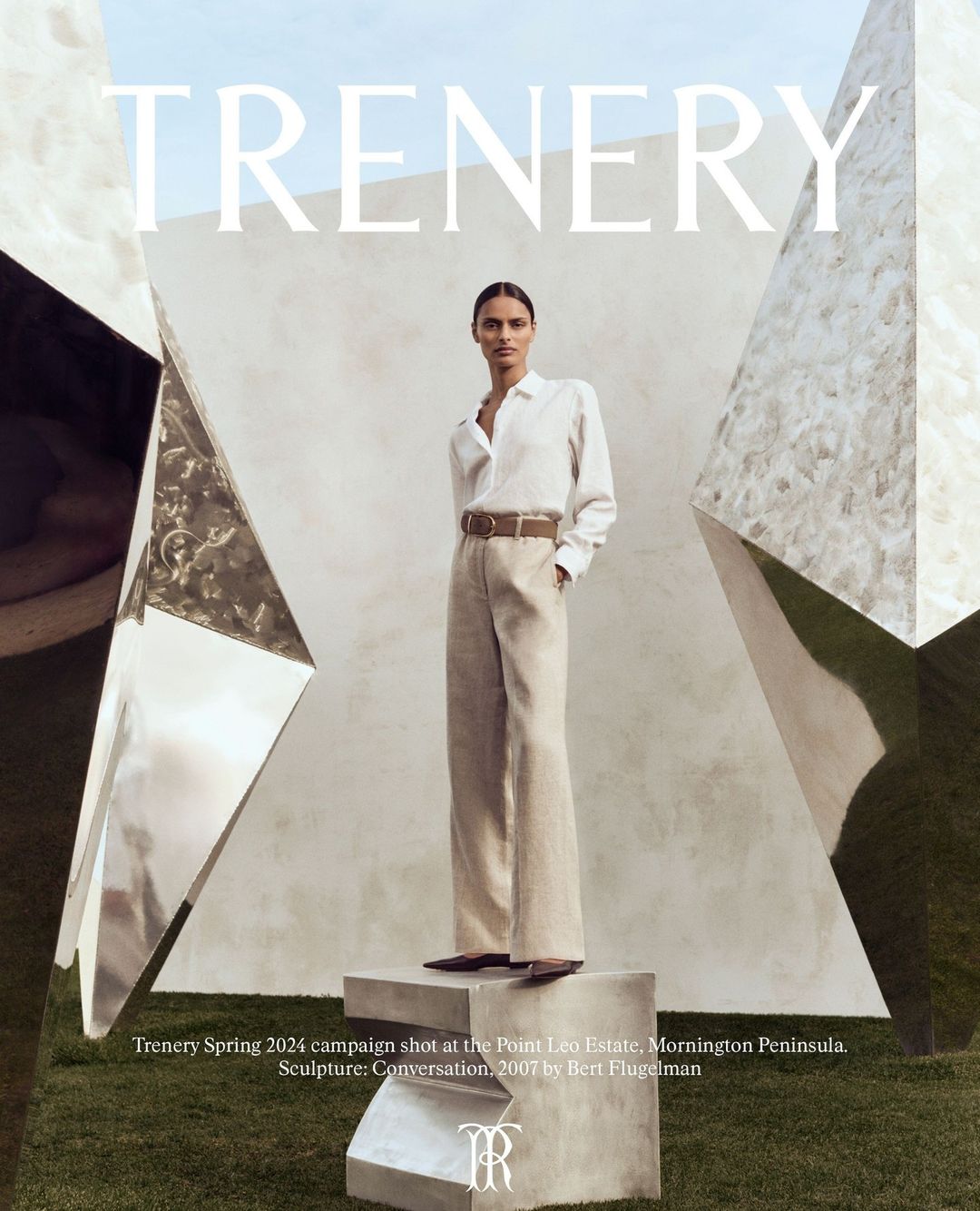 Fashion editorial featuring a woman in neutral clothing posing on a concrete block next to geometric sculpture in an outdoor setting.