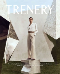 Model standing on a concrete plinth wearing a white button-down shirt and beige trousers between large reflective sculptures