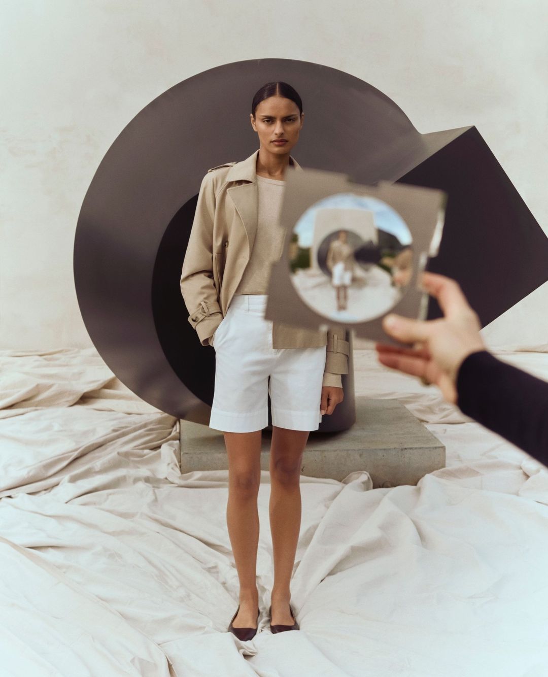 Fashion shot of a woman in shorts and trench coat posing in front of a gray geometric sculpture with a reflected scene. 