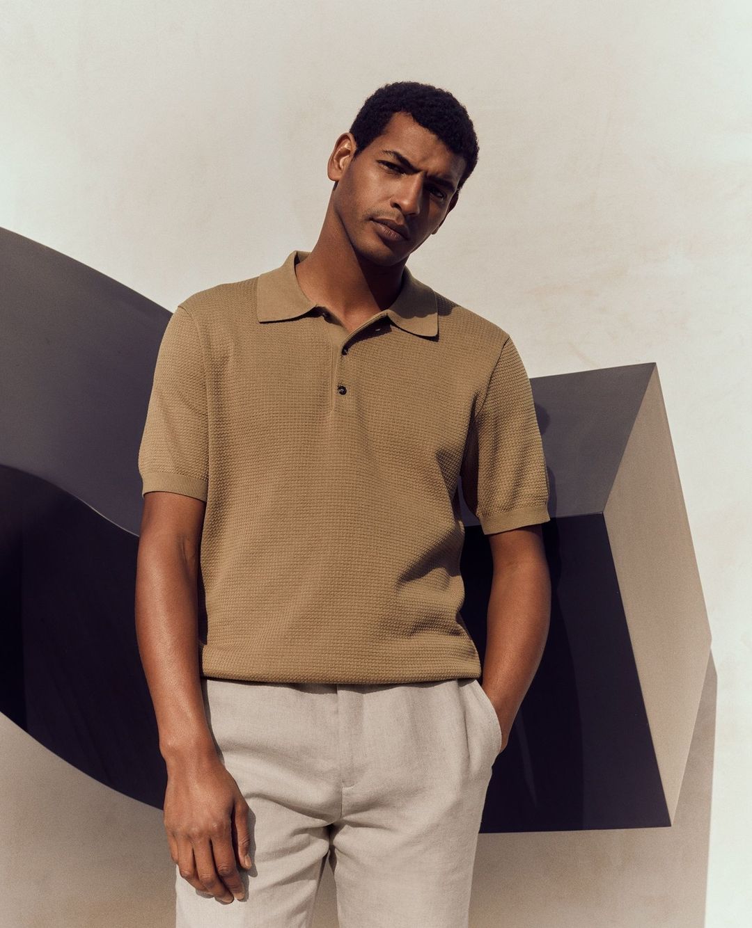 A stylish adult male wearing a tan polo shirt and light-colored slacks poses against a modern geometric backdrop indoors.