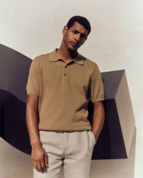 Man wearing a camel knit polo shirt and light grey trousers posing against a minimalist sculptural backdrop.