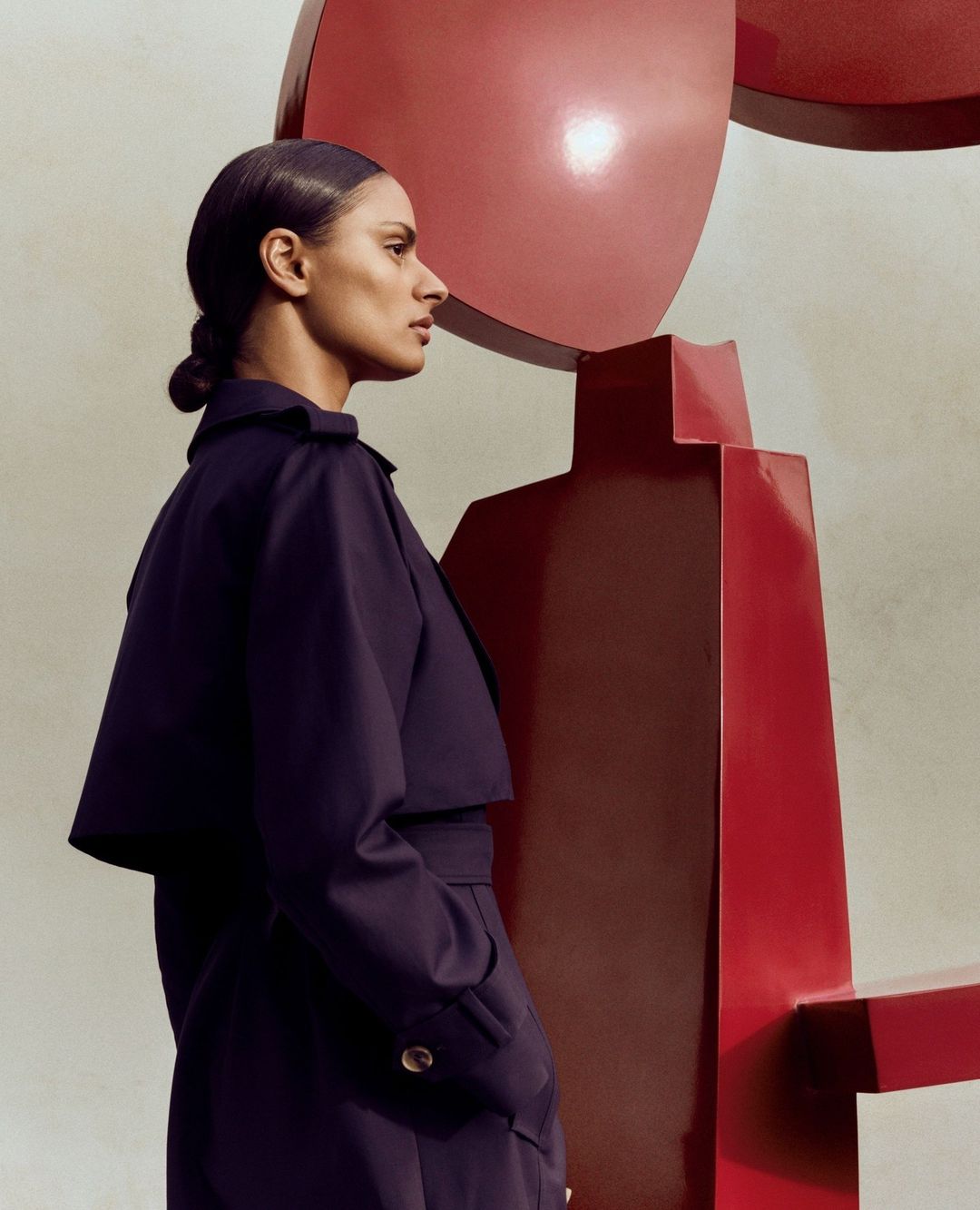 Profile view of an adult woman with dark hair in a navy outfit, standing next to a modern red sculpture against a neutral background.