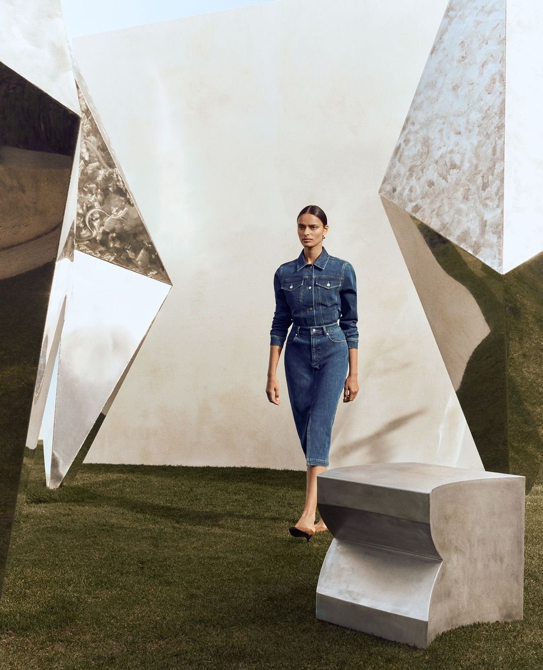 Fashion shot of a woman in a denim outfit, standing in a green space, framed by silver geometric art structures and a blocky seat.