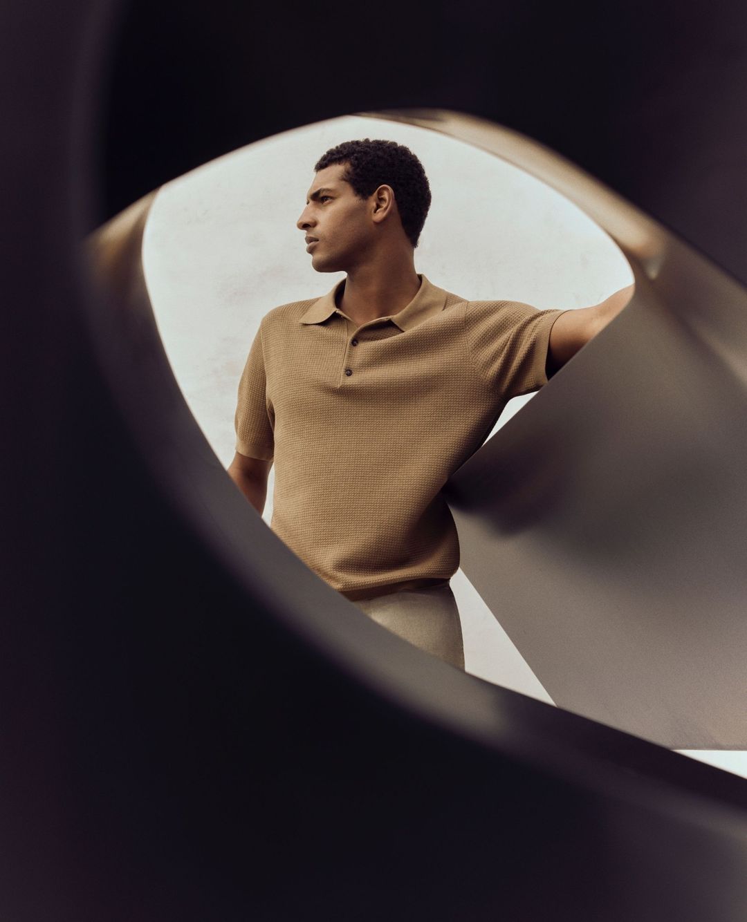 A man in a tan polo shirt poses with a dark gray abstract sculpture against a neutral background, captured in a medium shot.