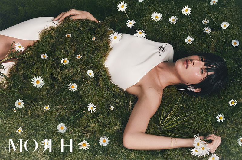 Brunette woman in white dress lying in grass covered in daisies. Fashion shot with grass textures and natural light in outdoor setting.