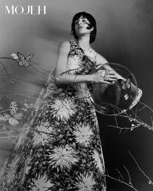 Black and white fashion portrait of a woman in a floral dress holding a glass orb with butterflies and standing against a dark background.