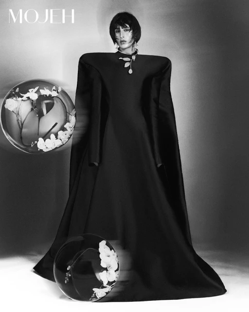 Black and white studio shot of a female fashion model in a black gown with wide shoulders, accessorized with a leaf pendant.