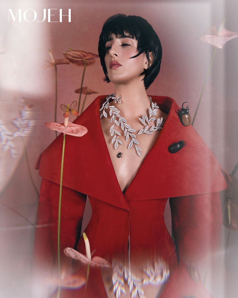 Woman in a red coat and jewelry stands in front of anthurium flowers in a studio with diffused lighting, striking a stylish pose.