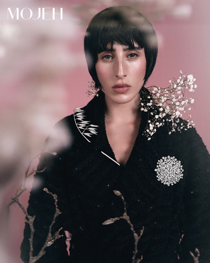 Close-up portrait of a woman with a bob haircut wearing a black jacket with white floral accents and a brooch against a pink background.