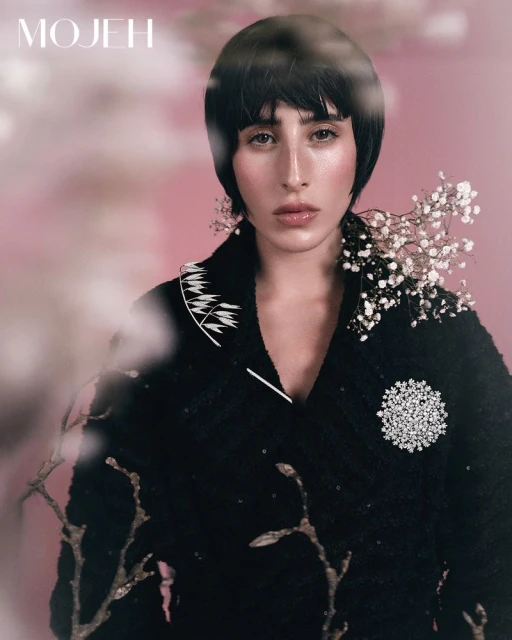 Close-up portrait of a woman with a bob haircut wearing a black jacket with white floral accents and a brooch against a pink background.