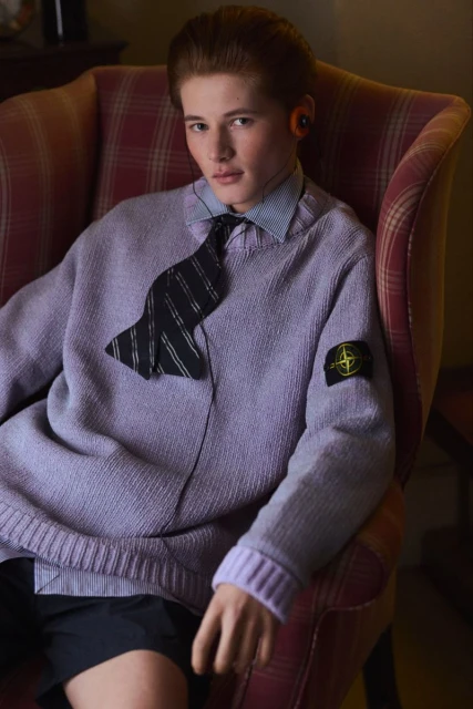 Man seated in a patterned armchair, wearing a lavender sweater, collared shirt, and tie; fashion portrait with muted indoor lighting.