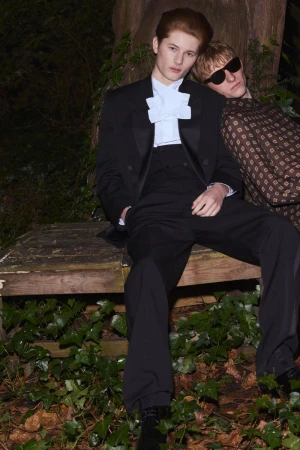 Two young men dressed in formal attire pose on a wooden structure outdoors, surrounded by lush greenery in a dimly lit environment.