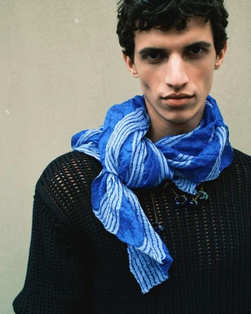 Close-up of a young adult male model wearing a blue patterned scarf and a black knitted sweater, posing against a neutral wall.