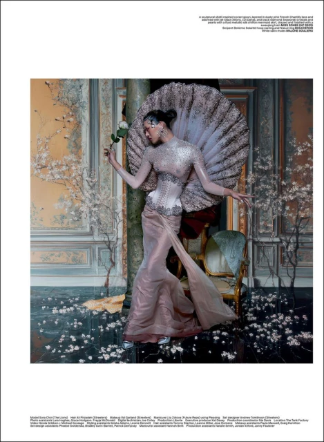 A woman in an elaborate sheer dress holds a white rose in an ornate room with faux cherry blossoms and marble floors.