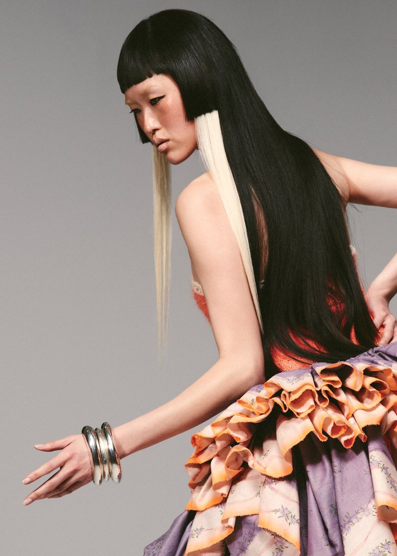 Asian woman with black and blonde straight hair poses in a light gray studio wearing silver bracelets and a purple ruffle dress.