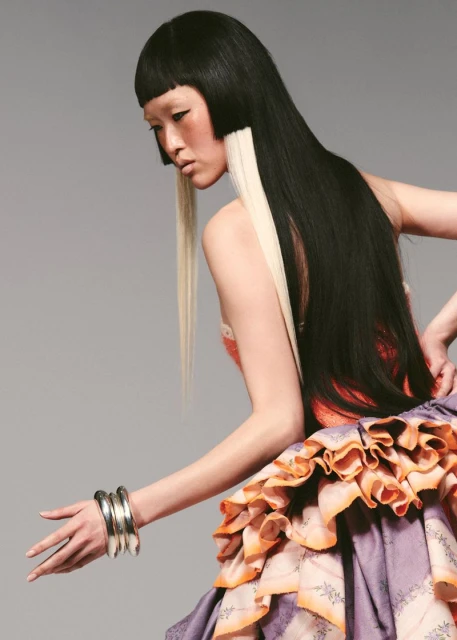 Asian woman with black and blonde straight hair poses in a light gray studio wearing silver bracelets and a purple ruffle dress.