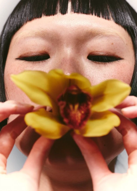 Close-up of an Asian woman with blunt bangs holding a yellow orchid flower in front of her face, shot in a beauty and fashion style.