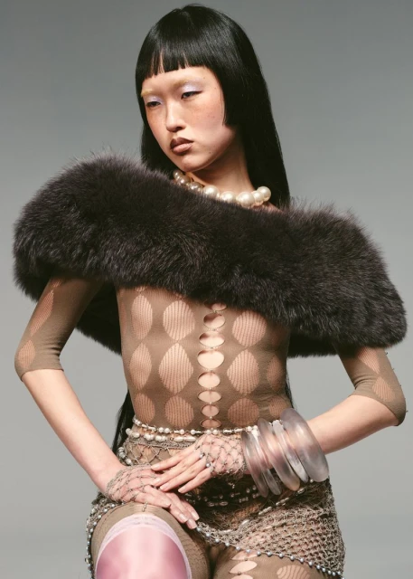Fashion shot of a young Asian woman wearing a patterned bodysuit, fur stole, pearls, and clear bracelets against a neutral backdrop.