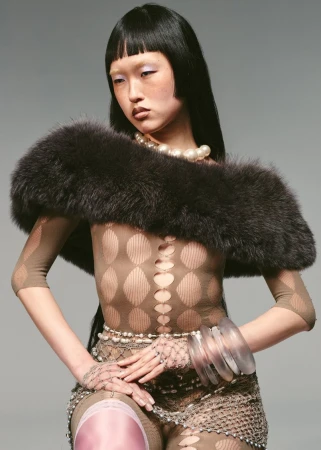 Fashion shot of a young Asian woman wearing a patterned bodysuit, fur stole, pearls, and clear bracelets against a neutral backdrop.