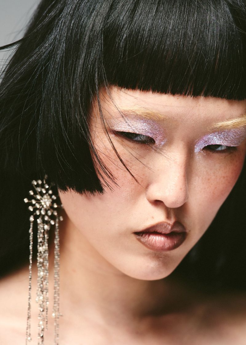 Headshot of a young adult woman with black hair, light purple eye makeup, and long, crystal chandelier earrings in a studio setting.