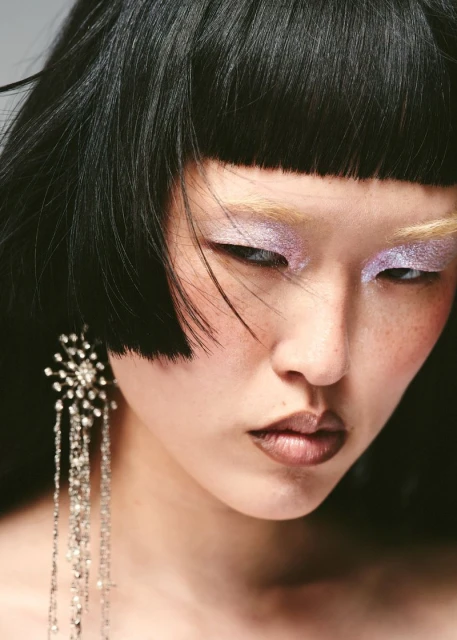 Headshot of a young adult woman with black hair, light purple eye makeup, and long, crystal chandelier earrings in a studio setting.