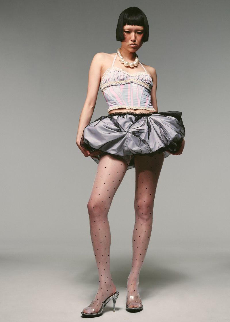 Full-length portrait of a young woman with a bob hairstyle, wearing a light-colored bustier top, black tutu skirt, and polka dot tights.