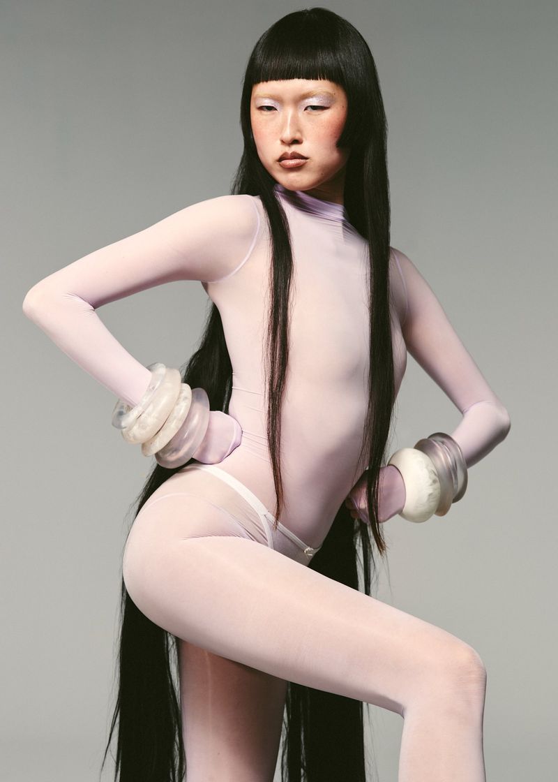 Asian woman in a light lavender bodysuit strikes a pose with exaggerated bangle bracelets in a studio setting. Full body shot.