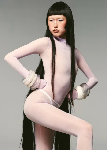Asian woman in a light lavender bodysuit strikes a pose with exaggerated bangle bracelets in a studio setting. Full body shot.