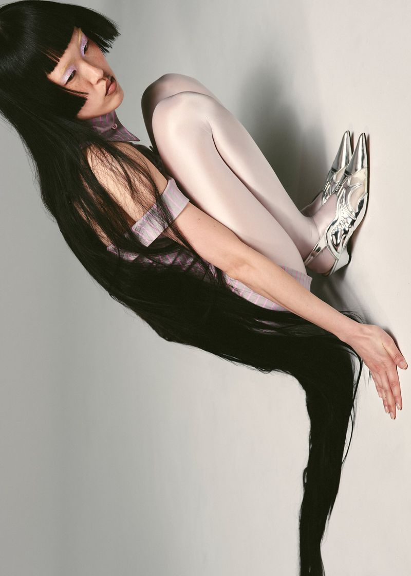 Fashion shot of a single woman in an unusual pose wearing metallic shoes and light purple clothing, sitting against a pale grey background.