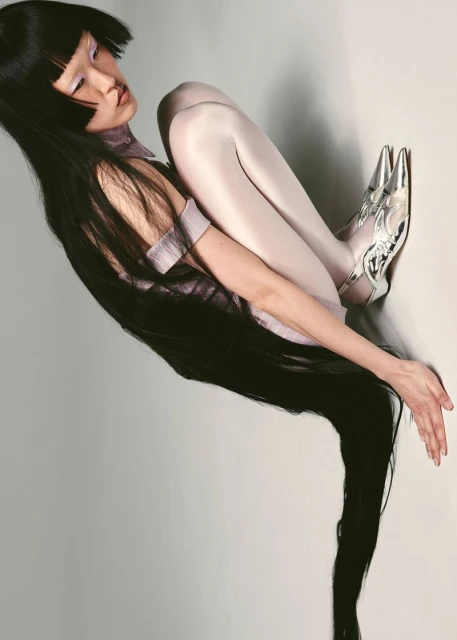 Fashion shot of a single woman in an unusual pose wearing metallic shoes and light purple clothing, sitting against a pale grey background.