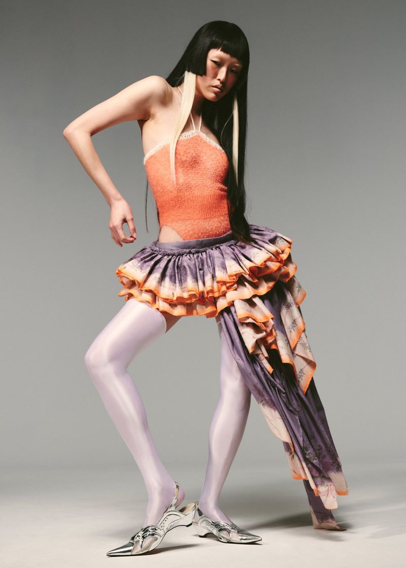 A slender woman with striking black and white hair poses in a studio wearing an orange halter top, layered ruffle skirt, and silver heels.