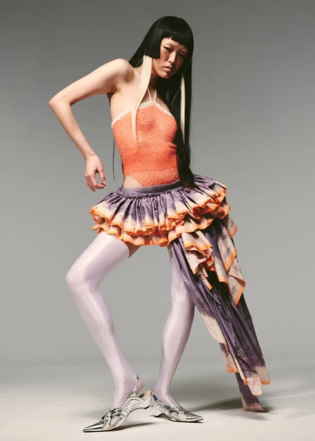 A slender woman with striking black and white hair poses in a studio wearing an orange halter top, layered ruffle skirt, and silver heels.