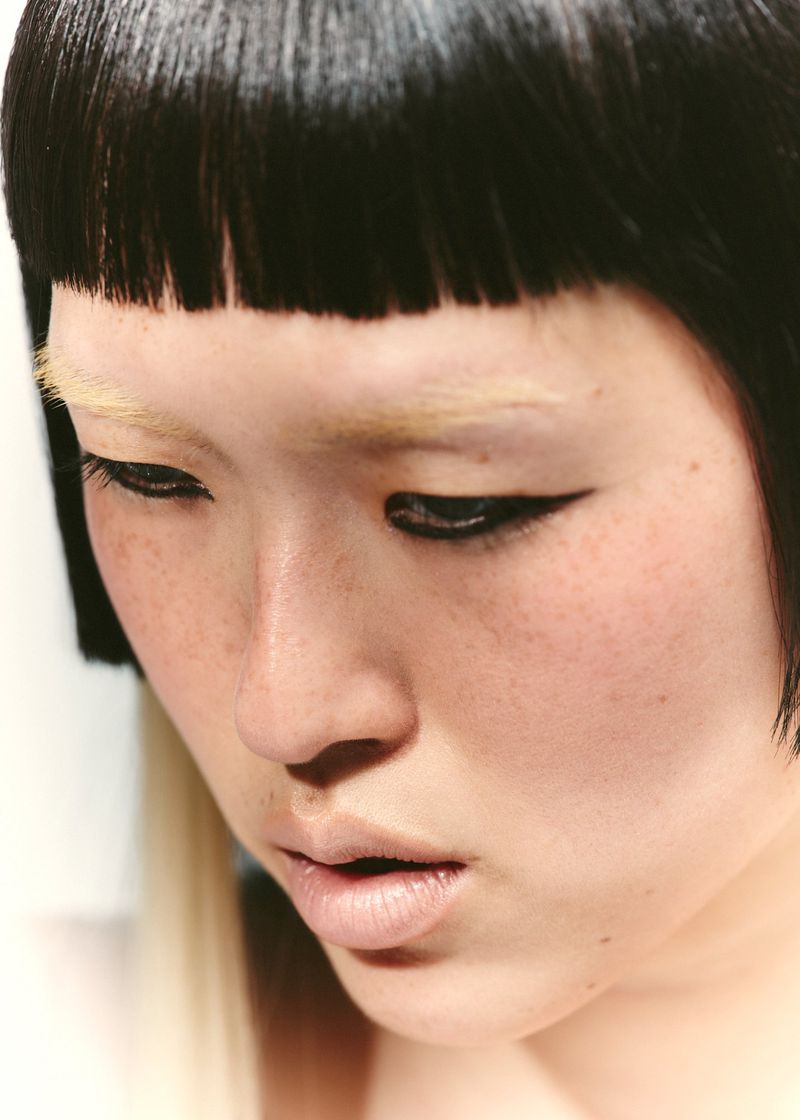Close-up portrait of an Asian adult female with fair skin, black bowl cut, winged eyeliner, and bleached eyebrows, looking down.