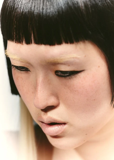 Close-up portrait of an Asian adult female with fair skin, black bowl cut, winged eyeliner, and bleached eyebrows, looking down.