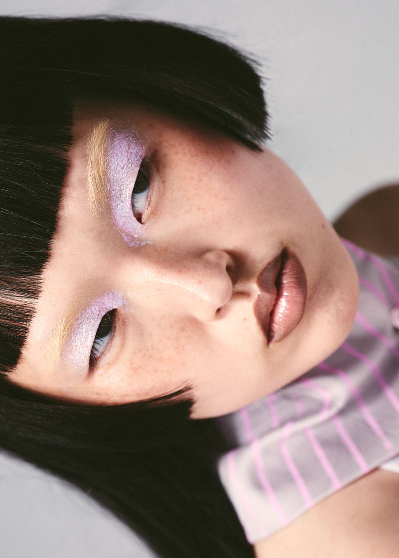 Close-up portrait of an Asian woman with dark hair, light purple eyeshadow, and glossy lips, wearing a pink and white striped shirt.
