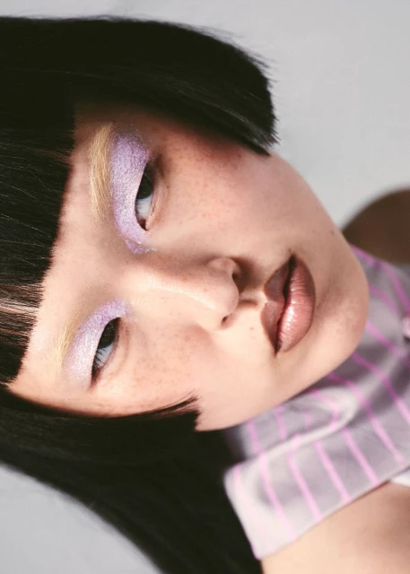 Close-up portrait of an Asian woman with dark hair, light purple eyeshadow, and glossy lips, wearing a pink and white striped shirt.