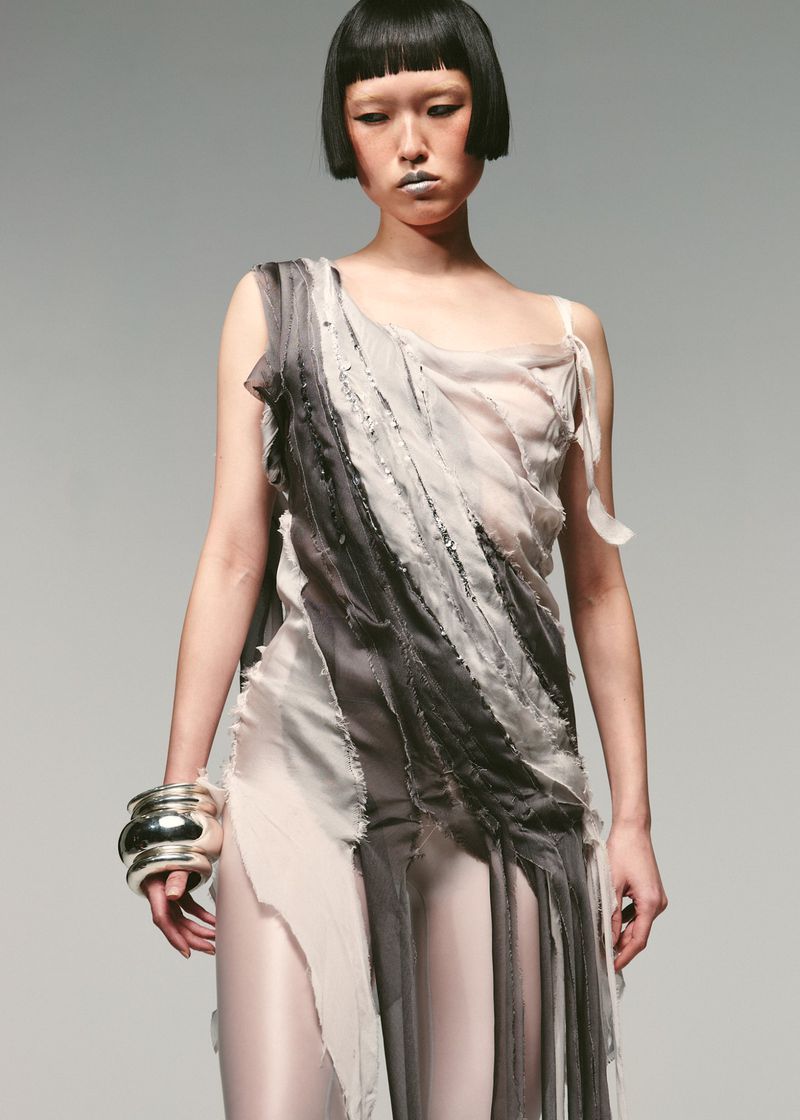 Elegant runway shot of a woman with blunt black hair in a flowing semi-sheer white and grey gown, posing against a light grey background.