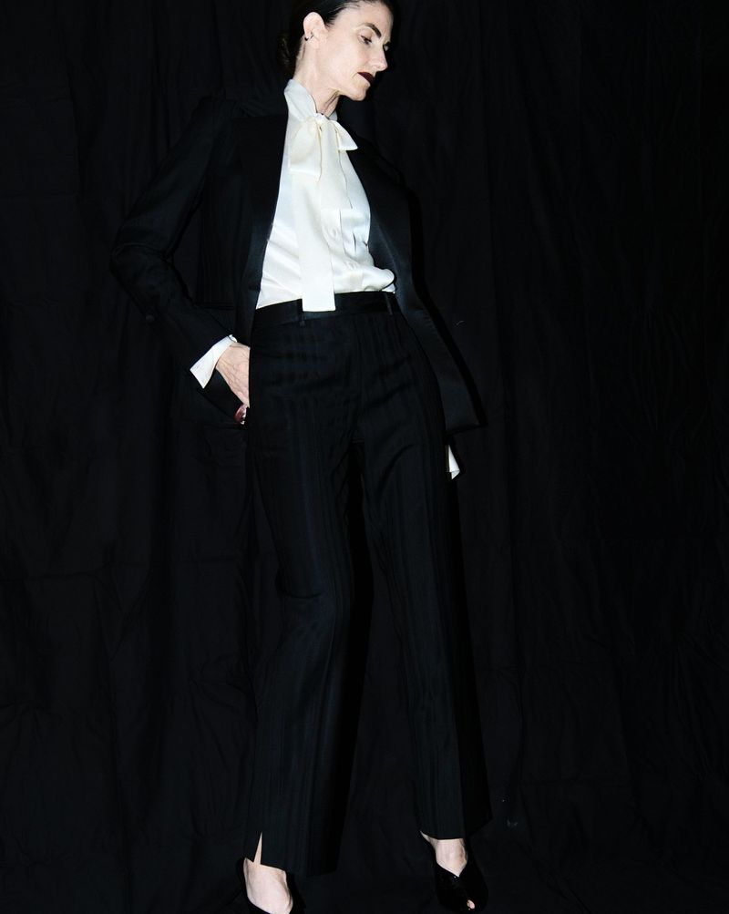 Woman in a black suit and white blouse poses in front of a dark backdrop, looking off to the side with a neutral expression.