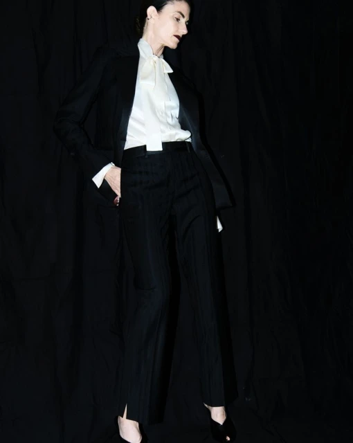 Woman in a black suit and white blouse poses in front of a dark backdrop, looking off to the side with a neutral expression.