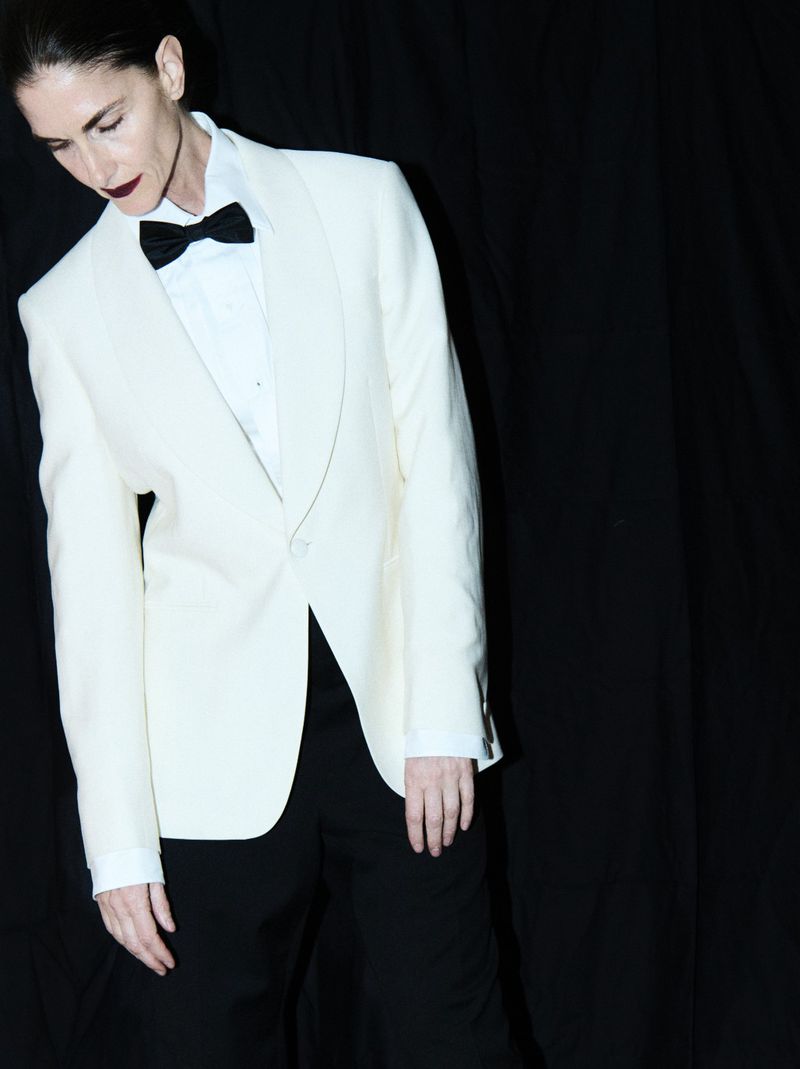 Elegant woman poses in a white tuxedo jacket, black bow tie, and black pants against a stark black backdrop, creating a striking contrast.